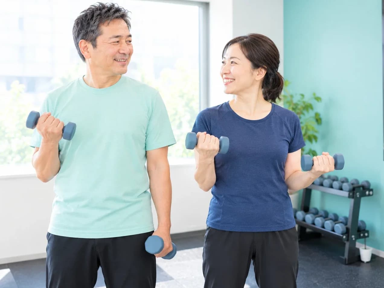 個室で並んでダンベルを持ち、顔を見合わせて笑う50代の夫婦