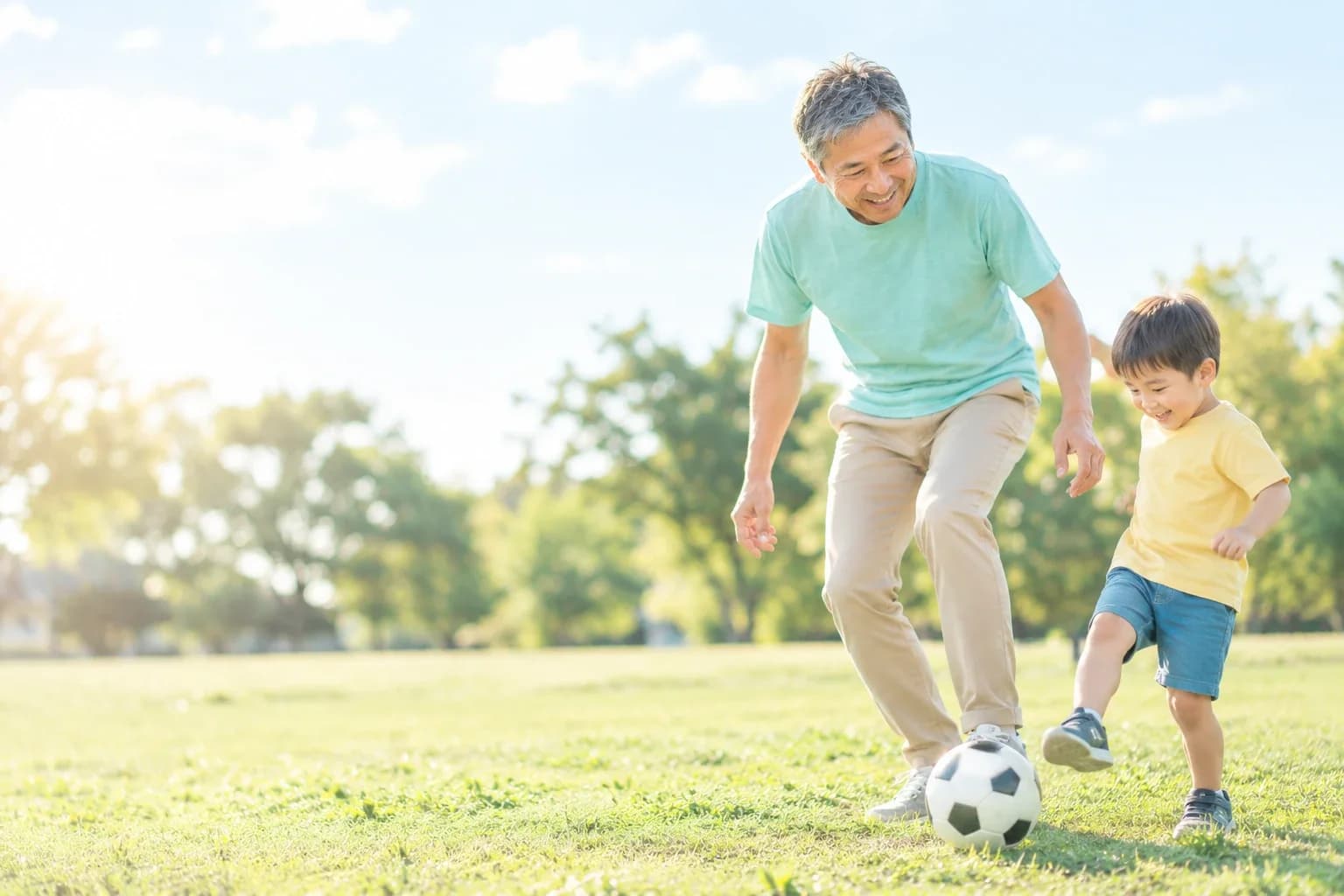 晴れた公園の芝生で、3歳くらいの孫とサッカーボールで遊ぶ男性